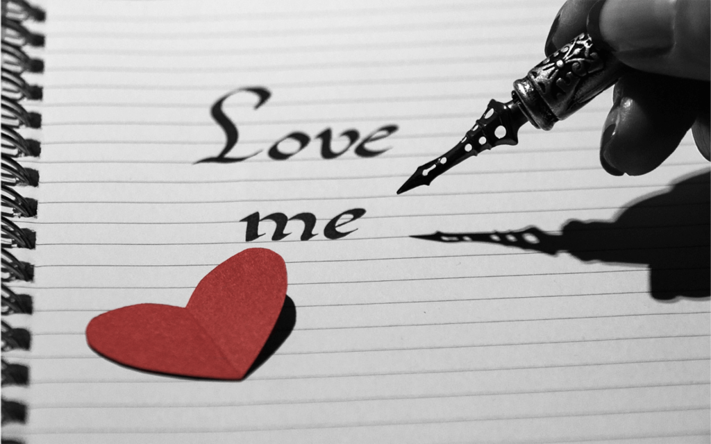 Note page with script saying "Love Me" and a cutout red paper heart and a calligraphy pen being held by a woman