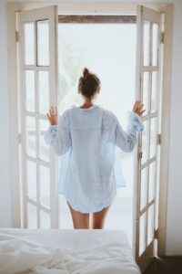 Woman with long light blue men's shirt opening french doors to a beautiful, sunny day.